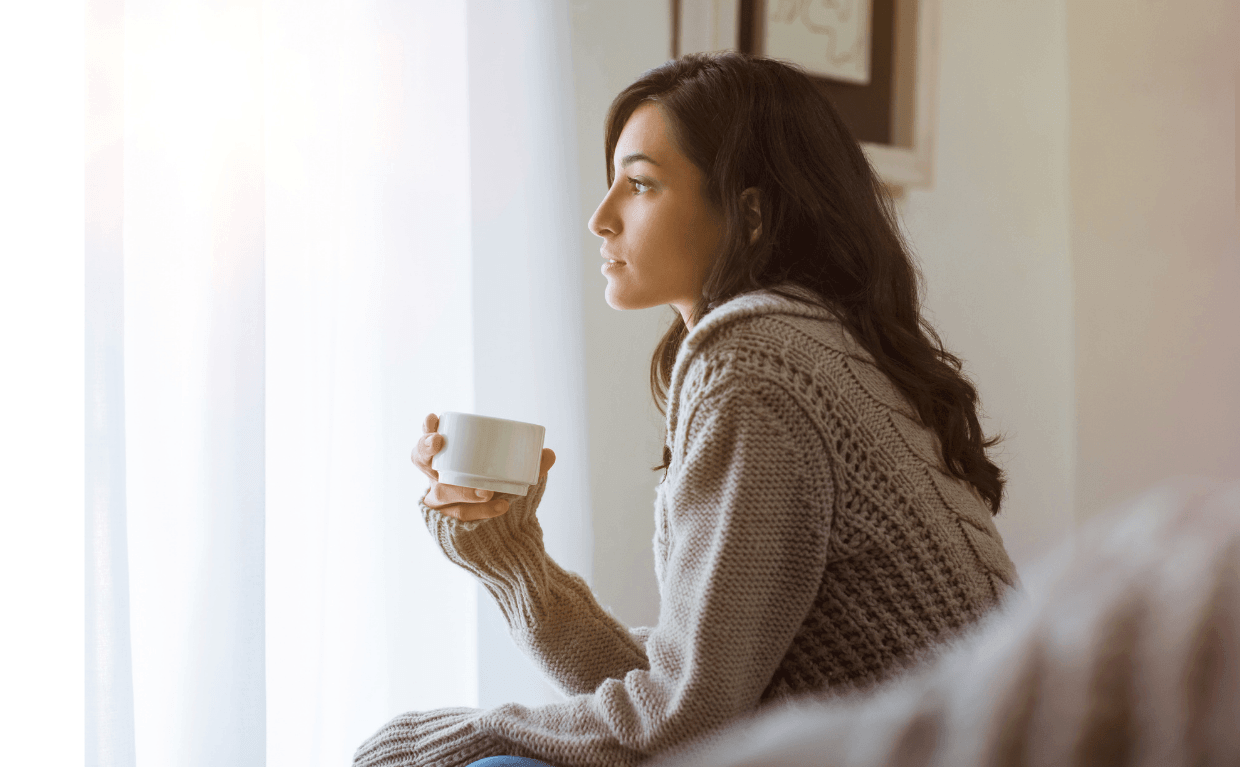 Frau mit Tasse in der Hand sieht nachdenklich aus dem Fenster